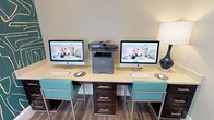 This business center is a compact yet fully functional workspace designed to meet a variety of professional needs. The room is anchored by a long, light-colored work surface accommodating two Apple iMac computers, each paired with a wireless keyboard and mouse, making simultaneous use straightforward and efficient. A Brother multifunction printer and scanner sits centrally on the desk, providing convenient print and copy access. Teal-accented bar-height chairs with sleek metal frames complement the desk's dark espresso storage drawers, lending a cohesive and modern aesthetic throughout.

One of the most striking design elements is the bold, large-scale abstract wallpaper in deep teal with gold linework, which covers select walls and adds a dramatic visual focal point to the otherwise neutral space. The remaining walls are finished in a soft, light gray, creating a balanced contrast. Wood-look flooring runs throughout, adding warmth underfoot.

Natural light enters through a window fitted with horizontal blinds, offering views of the surrounding area. A French glass door provides entry and maintains a sense of openness while visually connecting the business center to adjacent spaces. Ambient lighting from a decorative table lamp rounds out the well-considered atmosphere, making this center both practical and visually engaging.