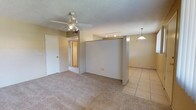 This single-floor residence offers a practical and comfortable layout designed for easy living. The main living area is finished with plush neutral carpeting, creating a warm and inviting atmosphere, while the adjoining dining or kitchen zone transitions to smooth tile flooring, providing a clear visual distinction between the two spaces. A half-wall partition elegantly separates the living and dining areas without closing off the open feel of the floor plan.

Overhead, a ceiling fan with integrated lighting serves the carpeted living space, offering both comfort and illumination. Track lighting is mounted along the partition wall, providing directional light to highlight key areas of the room. A hanging pendant light anchors the tiled dining area, adding a focused decorative touch. The textured popcorn ceiling adds a classic residential character throughout.

Neutral, light-toned walls keep the space feeling bright and versatile, ready to complement a variety of interior styles. A hallway visible from the living area leads to additional rooms, suggesting a well-connected and functional floor plan. A door on the far wall provides access to what appears to be a utility or storage space. Overall, the residence presents a clean, move-in-ready environment with a cohesive and understated aesthetic.