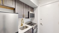 This efficiently designed 1-bedroom, 1-bathroom unit makes excellent use of its compact footprint across a single floor. The layout flows logically from the kitchen through to the living and sleeping areas, connected by a narrow hallway with wood-look flooring that runs throughout the main spaces.

The kitchen is thoughtfully appointed with dark gray shaker-style cabinetry accented by brushed gold hardware, white quartz countertops, and a classic white subway tile backsplash. Stainless steel appliances include a full gas range with overhead microwave, a compact refrigerator, and a deep single-basin sink fitted with a gold-finish pull-down faucet — all combining to create a polished, modern aesthetic in a small but functional space.

The bedroom is a generous portion of the overall unit, featuring hardwood-style flooring, neutral white walls, a double closet, and a window with a built-in air conditioning unit. A baseboard heating system runs along the perimeter, ensuring year-round comfort.

The bathroom presents a clean, contemporary design with floor-to-ceiling white subway tile surrounding the tub-shower combination. Matte black fixtures — including the showerhead, faucet, and drain — contrast elegantly against the bright white surfaces, while a white vanity with a sleek countertop and mirrored medicine cabinet complete the look.