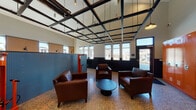 This single-floor facility at 4528 SW 39th St in Redmond, Oregon, is a thoughtfully designed amenity building offering a variety of functional spaces under soaring ceilings. The main lounge area features polished terrazzo flooring, a striking grid-pattern ceiling with suspended black steel framing, and abundant natural light streaming through large windows that frame views of the surrounding neighborhood. Rich brown leather seating arrangements centered around a round coffee table create a welcoming gathering space.

A dedicated bike maintenance zone includes a red upright bike repair station and a pegboard tool wall, positioned alongside the lounge for convenient access. Vibrant orange metal lockers provide secure personal storage nearby. The open work or activity area offers mobile worktables and a long wall-mounted desk beneath generous windows, making it a versatile multipurpose space.

The pet care wing is a standout feature, housing a dedicated grooming area with a utility sink set into dark navy cabinetry, a wall-mounted TV, and a sliding barn door. An adjacent kennel room contains rows of heavy-duty wire enclosures with polished floors and a narrow window for ventilation. Throughout, the two-tone black and white wall treatment, polished concrete floors, and industrial-modern detailing give the entire facility a cohesive, contemporary character.