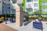 Located at 45 E Reed St in the heart of San Jose, California, this outdoor amenity space is a thoughtfully designed courtyard that serves as a communal gathering hub. The space is defined by wide, paver-lined walkways that guide visitors through lush, curated landscaping, including raised planter beds filled with ornamental grasses, flowering plants, and low-maintenance shrubs bordered by Corten steel and masonry retaining walls.

A standout feature of the courtyard is the open-air kitchen and grilling station, equipped with built-in BBQ grills, stone-topped counter surfaces, and bar-height seating — all set atop a warm wood deck platform ideal for casual outdoor dining. Adjacent to this, a comfortable lounge area with upholstered seating offers a relaxed setting for social gatherings.

Large decorative light poles are distributed throughout the space, providing illumination for evening use. A covered transitional area with full glass garage-style doors seamlessly connects the outdoor space to an interior room, offering flexible indoor-outdoor functionality with outdoor table-and-chair seating just outside.

The surrounding multi-story building facade features a bold, contemporary color palette of yellow, green, blue, and gray. Sweeping views of palm trees and the open San Jose skyline under abundant California sunshine complete the vibrant, resort-like atmosphere of this well-appointed outdoor amenity level.