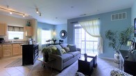 This single-floor residence offers a welcoming and well-designed open-concept layout that seamlessly connects the living, dining, and kitchen areas. The living room is a comfortable focal point, featuring plush carpeted flooring, a generous sofa accented with decorative pillows, and stylish dark wood furniture including a sleek coffee table and side tables. Soft blue-grey walls lend a calming, airy ambiance throughout the space. Natural light pours in generously through multiple windows dressed with sheer, flowing curtains, as well as a large sliding glass door that opens to a private balcony, offering an inviting connection to the outdoors. A decorative sunburst mirror adds a distinctive artistic touch to the living area wall. The dining area sits adjacent to the living room, furnished with a dark wood table and matching chairs, and is illuminated by a wall sconce that enhances the intimate atmosphere. The kitchen is positioned just beyond, featuring cabinetry in a warm wood tone and dark countertops visible along a functional bar-style divider. Track lighting on the ceiling provides additional illumination, complementing the abundant natural light. Overall, the space exudes a relaxed yet polished character, ideal for comfortable everyday living.