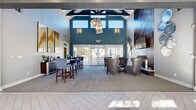 This well-appointed single-story clubhouse offers a thoughtfully designed layout ideal for both socializing and private meetings. The centerpiece is a dramatic great room with soaring vaulted ceilings accented by exposed dark beams and a bold teal feature wall. A striking cluster pendant light fixture draws the eye upward, while floor-to-ceiling glass doors flood the space with natural light and provide direct access to an outdoor patio. A tiled fireplace anchors one end of the lounge, surrounded by plush leather seating and circular decorative wall art, creating an inviting gathering spot. A coffee and refreshment bar with high-top seating lines one wall, finished with warm wood cabinetry and elegant abstract artwork above. The herringbone-patterned tile flooring in the entry transitions seamlessly to neutral carpet throughout the lounge area. A private meeting or conference room, enclosed by glass-panel sliding doors, features a round dark table with upholstered white chairs and a dramatic mountain landscape mural as a focal point. An adjacent lounge area offers comfortable blue leather chairs, light wood flooring, recessed lighting, crown molding, and large windows that bring in abundant natural light, completing this versatile and sophisticated community space.