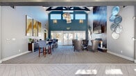This well-appointed single-story clubhouse offers a thoughtfully designed layout ideal for both socializing and private meetings. The centerpiece is a dramatic great room with soaring vaulted ceilings accented by exposed dark beams and a bold teal feature wall. A striking cluster pendant light fixture draws the eye upward, while floor-to-ceiling glass doors flood the space with natural light and provide direct access to an outdoor patio. A tiled fireplace anchors one end of the lounge, surrounded by plush leather seating and circular decorative wall art, creating an inviting gathering spot. A coffee and refreshment bar with high-top seating lines one wall, finished with warm wood cabinetry and elegant abstract artwork above. The herringbone-patterned tile flooring in the entry transitions seamlessly to neutral carpet throughout the lounge area. A private meeting or conference room, enclosed by glass-panel sliding doors, features a round dark table with upholstered white chairs and a dramatic mountain landscape mural as a focal point. An adjacent lounge area offers comfortable blue leather chairs, light wood flooring, recessed lighting, crown molding, and large windows that bring in abundant natural light, completing this versatile and sophisticated community space.