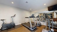 Located at 962 W Second St in Rialto, California, this single-room fitness center offers a well-organized and functional workout environment. The space is thoughtfully designed to maximize its compact footprint, accommodating a variety of cardio and strength-training equipment. Multiple treadmills are positioned along one wall, complemented by a stationary upright bike, an elliptical cross-trainer, and a multi-station cable strength machine with an adjustable bench — providing a comprehensive range of exercise options.

A large wall-mounted mirror spans a significant portion of the room, visually expanding the space while allowing users to monitor their form during workouts. The interior features neutral-toned walls, recessed ceiling lighting that ensures even illumination throughout, and carpeted flooring with rubber exercise mats placed beneath the equipment for stability and protection. A wall-mounted television is also present, adding convenience for users during cardio sessions.

Natural light enters through a windowed entry door flanked by sidelights, offering views of an outdoor common area with greenery, giving the space a welcoming atmosphere. The clean, straightforward design keeps the focus on functionality, making this fitness center a practical and accessible amenity for residents of the surrounding community.