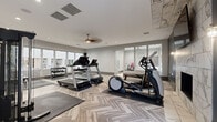 Located at Overland Station in Overland Park, Kansas, this single-floor fitness center is thoughtfully designed to deliver a comprehensive workout experience across multiple distinct zones. The main cardio area showcases striking herringbone-patterned hardwood-style flooring, neutral gray walls, and large floor-to-ceiling windows that flood the space with natural light while offering views of the surrounding community. A ceiling fan and recessed lighting ensure comfort during workouts. This area is equipped with treadmills, ellipticals, and a multi-use cable machine, complemented by a full dumbbell rack and weight benches for strength training. A marble-accented accent wall with a wall-mounted flat-screen TV adds a refined, upscale touch. A dedicated cycling studio features polished marble-style tile flooring, roller-shade windows, built-in shelving, a mini refrigerator, and a mounted TV — creating an immersive spin class environment. Floor-to-ceiling mirrors throughout amplify the sense of space and allow for form checks during exercise. A separate flexible zone provides storage for yoga mats, resistance bands, stability balls, and other functional fitness accessories, making the facility well-suited for a variety of training styles. The overall ambiance is modern, clean, and inviting.