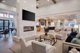 Located at 74 Hauppauge Rd in Commack, NY, this elegantly designed single-floor community space offers a generous, open layout that seamlessly connects multiple functional areas. The living room serves as the heart of the space, anchored by a dramatic floor-to-ceiling marble fireplace column with a built-in linear gas fireplace and a large wall-mounted TV. Soaring ceilings with exposed wood beams, herringbone light wood flooring, and abundant natural light from oversized windows and glass doors create a bright, airy atmosphere throughout. A dedicated dining area accommodates generous seating arrangements beneath a coffered ceiling with a stylish brass chandelier, while wainscoting and arched windows add classic architectural detail. The bar area features a large white quartz countertop island with bar stool seating, warm wood cabinetry, a beverage station, and a dramatic cluster of pendant lights overhead. The kitchen is outfitted with oak cabinetry, a tile backsplash, white countertops, and stainless steel appliances, complemented by natural light from plantation-shuttered windows. A game room with a pool table, casual seating, and nautical-themed artwork rounds out the space, offering a relaxed yet refined retreat. The overall ambiance is sophisticated, coastal-inspired, and welcoming.