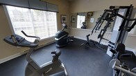 This fitness room is thoughtfully arranged to accommodate a variety of workout needs within a single-floor space. The room features large windows fitted with horizontal blinds, allowing natural light to fill the area while maintaining privacy. A full-length wall mirror extends along one side, visually expanding the room and allowing users to monitor their form during exercise. The dark, rubberized flooring provides durability and cushioning underfoot, making it well-suited for active use. Equipment includes a stationary elliptical trainer, a stationary exercise bike, and a multi-function cable strength training machine, offering options for both cardiovascular and resistance training. Framed artwork is displayed on the neutral-toned walls, adding a modest decorative touch to the otherwise functional space. A baseboard heating unit runs along the exterior wall beneath the windows, ensuring year-round comfort. The layout is efficient, with equipment positioned to allow reasonable movement between stations. Overall, the room presents a practical and well-lit environment designed to support a range of fitness routines.
