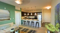 This single-floor space showcases a well-designed open-concept layout that seamlessly integrates the kitchen and living area. The kitchen is a standout feature, anchored by a generous island with light granite-style countertops and seating for four, making it ideal for casual dining and entertaining. Dark espresso cabinetry lines the walls, providing ample storage and creating a bold contrast against the lighter countertops and cream-toned walls. Stainless steel appliances, including a refrigerator and microwave, add a modern, polished touch. Above the island, a trio of sleek pendant lights offers focused task lighting, while a flush-mount ceiling fixture illuminates the living area. An accent wall painted in a rich green tone adds a pop of color and visual depth to the space. The living area flows naturally from the kitchen, featuring plush neutral seating and a patterned area rug that defines the zone. Light-toned flooring and carpet contribute to the airy feel throughout. Interior doors are finished in clean white, complementing the overall neutral palette. The combined layout promotes an inviting, functional ambiance suited for both everyday living and hosting.