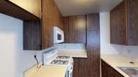 The kitchen in this single-floor space is defined by its extensive warm-toned wood cabinetry, which extends from counter height to the ceiling, maximizing vertical storage throughout the room. The cabinetry features a rich walnut-style wood grain finish that lends the space a classic, traditional character. A white gas range with four burners serves as the cooking centerpiece, complemented by a matching over-the-range microwave mounted directly above, providing both ventilation and additional cooking convenience. Light-colored countertops offer a clean contrast against the darker cabinetry, creating visual balance within the kitchen. The layout follows an L-shaped configuration, efficiently utilizing the available space while allowing for a practical workflow between the cooking and preparation areas. Additional cabinetry is positioned along the adjacent wall, providing further storage capacity. The textured ceiling adds a subtle architectural detail consistent with the overall design of the space. The kitchen is well-equipped for everyday cooking needs, with its combination of ample cabinetry, functional appliances, and efficient use of the available footprint making it a practical and straightforward culinary space.