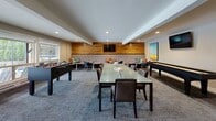 This thoughtfully designed single-floor amenity space serves as a versatile community lounge and recreation room, combining comfort, style, and entertainment in one open layout. The interior is anchored by a striking textured wood accent wall, which provides a warm, organic contrast to the neutral tones of the surrounding walls and recessed lighting overhead.

The lounge area features a generously sized tufted banquette with colorful accent pillows, complemented by contemporary accent chairs and small round tables — ideal for casual conversation or relaxing. A wall-mounted flat-screen television above the accent wall adds a media-viewing focal point to this seating zone.

At the center of the space, a long glass-topped dining table surrounded by leather upholstered chairs serves as a versatile gathering surface. Colorful horizon-inspired artwork adorns the walls, adding vibrant visual interest throughout.

The game area is well-equipped with a foosball table and a shuffleboard table, offering recreational options for residents and guests alike. Large windows along one wall flood the space with natural light and provide views of an outdoor pool area, seamlessly connecting the interior to the exterior environment.

A separate room, accessible through a glass-paneled door, features warm wood flooring and mirrored walls, functioning as a fitness or flex studio. The patterned carpet throughout the main space ties all zones together, contributing to a cohesive and inviting ambiance.
