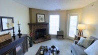 This single-floor residence offers a warm and inviting atmosphere anchored by a standout brick fireplace in the living room. The fireplace, complete with a dark wood mantel and decorative hearth, serves as the focal point of the space and adds a timeless, classic character. Neutral-toned walls complement the textured ceiling and plush wall-to-wall carpeting, creating a comfortable and cohesive aesthetic throughout. The living area is generously illuminated by natural light streaming through multiple windows, enhancing the open and airy feel of the room. A glass-paneled door provides additional light and access to the exterior. The space is thoughtfully arranged to balance functionality and comfort, with room for media storage, seating, and decorative accents. Subtle botanical touches, including framed floral artwork above the mantel and potted plants, add a natural warmth to the interior. The overall ambiance is relaxed and homey, making it well-suited for both everyday living and casual entertaining. The single-floor layout ensures ease of movement and accessibility throughout the home.