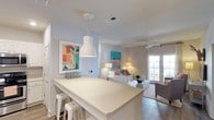 The Dozier property is a single-story space designed with an open-concept layout that connects the kitchen and living areas in a cohesive, flowing arrangement. The kitchen features a spacious peninsula counter with light stone-look surfaces, offering casual seating with white metal bar stools underneath. A white dome pendant light hangs above the counter, providing focused task lighting that complements the clean, contemporary aesthetic.

The transition into the living area is seamless, where hardwood-style flooring anchors the space. A ceiling fan with a light fixture is centrally positioned, promoting airflow and comfort throughout. French doors along the far wall allow generous natural light to filter in, enhancing the airy, welcoming ambiance. Neutral wall tones in soft gray and white serve as a backdrop to colorful framed artwork, adding visual interest without overwhelming the space.

Overall, the open layout, natural light, and modern fixtures combine to create a bright, functional, and comfortable living environment suited for everyday use.