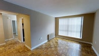 This single-floor space offers a clean and functional layout with a timeless aesthetic. The main living area immediately draws attention with its classic parquet wood flooring, arranged in a distinctive geometric pattern that adds warmth and character to the room. A large picture window fitted with vertical blinds allows an abundance of natural light to fill the space, creating a bright and airy atmosphere. A traditional radiator sits beneath the window, providing efficient heating. The neutral gray and warm beige tones on the walls complement the natural wood tones of the floor, contributing to a calm and understated ambiance. White baseboards provide a crisp contrast throughout the room. An open doorway leads from the main living area into an adjoining room, suggesting a connected and accessible floor plan that encourages easy movement between spaces. The overall design is straightforward and practical, with a focus on functionality while retaining the charm of classic architectural details such as the parquet flooring. The space presents a solid foundation well-suited for a variety of uses and interior styling preferences.