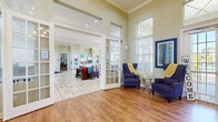 Located at 1222–1226 Floral Springs Blvd in Port Orange, Florida, this single-story community clubhouse is designed to provide residents with a warm, functional gathering space. The building's architecture is characterized by soaring ceilings with transom windows that draw in abundant natural light throughout, creating an open and airy atmosphere across all areas.

The entrance foyer greets visitors with warm hardwood-style flooring, a pair of navy blue armchairs, and large French doors that open to the outdoors. A curved wood reception desk serves as the central hub, lending the space a polished, professional feel. Tall clerestory windows above the reception area amplify the sense of height and openness.

Beyond the entry, the main gathering area features tile flooring and transitions seamlessly into multiple lounging zones, including a sunlit corner sitting room lined with large windows offering views of the surrounding landscape. A dedicated coffee bar area is equipped with granite countertops, white cabinetry, and built-in appliances, providing a comfortable refreshment station.

A business center with a built-in desk, computer workstation, and bistro-height seating offers a practical workspace. French doors throughout the clubhouse connect the various zones while maintaining an open, flowing layout. Crown molding and soft neutral wall tones complete the polished, community-oriented ambiance.