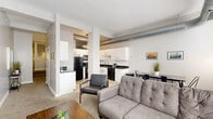 Located at 1211 W 9th St in Cleveland's vibrant Ohio City neighborhood, this two-bedroom, one-bathroom unit offers a well-designed single-floor layout with a blend of industrial character and modern comfort. The open-concept living and kitchen area serves as the heart of the unit, where exposed cylindrical ductwork adds an authentic loft aesthetic to the bright, neutral space. The kitchen features white cabinetry, dark granite countertops, black appliances, and light hardwood-style flooring, flowing seamlessly into the carpeted living area with large windows that invite natural light. A dedicated dining area sits adjacent to the kitchen. A carpeted hallway connects the main living space to the private quarters, lined with framing for artwork and illuminated by a sidelight window. The primary bedroom offers a window with drapes, exposed ductwork details, and comfortable proportions. The second bedroom provides additional space with a window, carpet flooring, and room for a workspace. The full bathroom includes a tub-shower combination with white surrounds and built-in overhead cabinetry. Three closets provide ample storage throughout, and an in-unit laundry area adds everyday convenience. The unit reflects a clean, contemporary aesthetic with industrial undertones throughout.