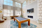 Located at 4111 N Drinkwater Blvd in Scottsdale, Arizona, this single-floor amenity lounge is a well-appointed gathering space designed with a modern, resort-inspired aesthetic. The room is defined by its dramatic double-height ceilings and a generous wall of floor-to-ceiling windows dressed in sheer linen curtains, flooding the interior with natural light and offering views of the surrounding outdoor greenery.

Light-toned hardwood-style flooring runs throughout, complementing the bright, airy atmosphere. A bold geometric diamond-patterned accent wall in neutral tones anchors the main living area and serves as a backdrop for a wall-mounted flat-screen television and a sleek built-in linear fireplace below. Flanking the feature wall are rattan hanging egg chairs with tropical print cushions and gold wall sconces that add warmth to the space.

A full-size pool table centered on a palm-leaf patterned area rug serves as the focal point of the room, while a shuffleboard table positioned near the windows offers additional recreation. A seating nook near the entry features woven rattan chairs beneath warm pendant lighting, complemented by botanical wallpaper accents. A kitchenette area with bar-height seating and a decorative laser-cut palm frond panel adds both function and artistic detail, completing this vibrant and inviting communal lounge.