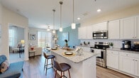 This single-floor residence is thoughtfully designed with a seamless open-concept layout connecting the kitchen, dining, and living areas. The kitchen serves as a focal point, showcasing white shaker cabinetry, granite countertop surfaces with subtle veining, a classic white subway tile backsplash, and a full suite of stainless steel appliances including a range, microwave, and refrigerator. A central island with pendant lighting overhead provides bar seating for casual dining and additional workspace. Wood-style flooring runs throughout the main living areas, lending warmth and continuity to the space.

The living area extends naturally from the kitchen, featuring a ceiling fan and access to natural light through a sliding glass door. A soft blue accent wall adds a distinctive visual element to the shared space.

The bedroom is carpeted for comfort and features a ceiling fan, multiple windows with blinds that allow ample natural light, and enough space for a full furniture arrangement including a desk area.

The bathroom is finished with a white vanity offering generous counter and cabinet storage, a large mirror, wood-style flooring, and a separate room housing the toilet and shower-tub combination, providing a practical and clean layout throughout.