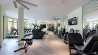 Located at 15705 SW 127th Ave in Miami, FL, this fitness center offers a generously sized, single-floor exercise facility designed to support a comprehensive range of workout routines. The space is bathed in natural light, thanks to floor-to-ceiling windows and glass walls that frame lush, tropical greenery outside — a fitting reflection of South Florida's vibrant landscape.

The interior is thoughtfully divided into distinct workout zones. A dedicated cardio area lines one side of the facility, equipped with a row of treadmills and stair climbers positioned to take advantage of the outdoor views. Large ceiling fans overhead provide airflow and contribute to a comfortable training environment. The light wood flooring throughout lends a clean, modern aesthetic to the space.

A strength training zone features power racks with Olympic barbells, a cable/pulley machine, adjustable benches, and additional resistance equipment, all positioned on rubber-mat flooring for safety and stability. Wall-mounted flat-screen televisions are integrated throughout for entertainment during workouts.

Glass partition walls with large aluminum-framed doors elegantly separate functional zones while maintaining a sense of openness. One section includes a glass roll-up garage door that opens directly to the outdoors, creating an indoor-outdoor training experience. The bright, airy ambiance, modern finishes, and diverse equipment selection make this facility a well-rounded and inviting fitness destination.
