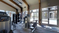 This well-appointed fitness center combines contemporary design with functional amenities, creating an inviting environment for exercise and wellness. The space is defined by soaring ceilings accented with prominent dark wood beams, lending a warm, architectural character that contrasts beautifully with the clean, modern finishes throughout.

Large floor-to-ceiling glass doors and expansive windows line the exterior wall, bathing the gym in abundant natural light while offering open views of the outdoor pool area and the surrounding residential community beyond. This seamless connection to the outdoors enhances the overall sense of space and airiness within the facility.

The fitness floor is outfitted with a comprehensive selection of equipment to support a variety of workout routines. Strength training is well-represented, with a cable machine, a seated resistance machine, and a full rack of dumbbells in graduated sizes. Functional training is accommodated with a collection of medicine balls and stability balls neatly organized on dedicated racks. The flooring features a durable, multi-tone carpet tile that defines the workout zones while providing comfort underfoot. Exposed ductwork on the ceiling adds an industrial edge that complements the overall modern-rustic aesthetic of the space.