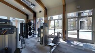 This well-appointed fitness center combines contemporary design with functional amenities, creating an inviting environment for exercise and wellness. The space is defined by soaring ceilings accented with prominent dark wood beams, lending a warm, architectural character that contrasts beautifully with the clean, modern finishes throughout.

Large floor-to-ceiling glass doors and expansive windows line the exterior wall, bathing the gym in abundant natural light while offering open views of the outdoor pool area and the surrounding residential community beyond. This seamless connection to the outdoors enhances the overall sense of space and airiness within the facility.

The fitness floor is outfitted with a comprehensive selection of equipment to support a variety of workout routines. Strength training is well-represented, with a cable machine, a seated resistance machine, and a full rack of dumbbells in graduated sizes. Functional training is accommodated with a collection of medicine balls and stability balls neatly organized on dedicated racks. The flooring features a durable, multi-tone carpet tile that defines the workout zones while providing comfort underfoot. Exposed ductwork on the ceiling adds an industrial edge that complements the overall modern-rustic aesthetic of the space.