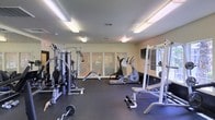 This single-floor fitness space is designed to support a comprehensive workout experience. The room is finished with dark rubber flooring that provides durability and cushioning underfoot, making it suitable for a range of exercise activities. Overhead, bright fluorescent panel lighting ensures the space is well-illuminated throughout.

The room houses a notable selection of equipment, including a multi-station cable and resistance machine, a power rack suitable for free weight training, a barbell with weight plates, and a bench for strength exercises. For cardiovascular training, an elliptical machine is positioned centrally within the space.

A flat-screen television is mounted on one wall, offering entertainment during workouts. Along the back wall, a set of large windows with horizontal blinds allows natural light to filter into the room while maintaining privacy. The walls are finished in a neutral light tone, contributing to a clean and functional atmosphere. The layout provides ample floor space between equipment, allowing for comfortable movement and use of multiple stations simultaneously.