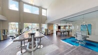 Located on Bruce B Downs Boulevard in Tampa, Florida, this single-floor amenity center is designed with a contemporary aesthetic and an impressive range of community features. The centerpiece is a grand clubroom with soaring double-height ceilings and floor-to-ceiling windows that flood the space with natural light and offer direct views of the outdoor pool area. A granite-topped bar with modern swivel stools anchors the lounge, while varied seating zones create flexible gathering spaces throughout. A glass-enclosed game room with a billiards table adds an entertaining focal point. An adjacent lounge nook features a curved, mosaic tile accent wall with pendant lighting, dark wood cabinetry, and a built-in kitchenette with stainless steel appliances. The expansive fitness center is outfitted with a full array of cardio machines, including treadmills positioned along large windows overlooking lush landscaping, as well as strength training equipment and open floor space. A glass-walled group fitness studio with hardwood flooring, mirrored walls, and exercise equipment provides a dedicated workout environment. A striking steel spiral staircase serves as a sculptural architectural element within the fitness area. Practical touches include a dual-basin water fountain station set against wood-panel walls with integrated storage.