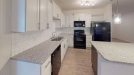 This single-story unit presents a thoughtfully designed layout with a consistent neutral color palette of soft whites and warm beiges throughout. The kitchen serves as a standout feature, showcasing white shaker-style cabinetry with brushed nickel hardware, granite countertops, a classic subway tile backsplash, and a central island with contrasting dark cabinetry. Black appliances, including a refrigerator, range, microwave, and dishwasher, complete the space. Light wood-style luxury vinyl plank flooring runs through the kitchen and living areas, while plush carpet covers the bedrooms.

The living area is bright and open, benefiting from natural light through window blinds and a ceiling fan overhead. Bedrooms are generously sized, featuring carpeted floors, two-panel gray doors, and closets with bi-fold doors and built-in rods for storage. The primary bathroom includes a frameless sliding glass shower enclosure, while a secondary bathroom offers a full soaking tub and shower combo with built-in shelving. Recessed lighting, ceiling fans, and clean white walls contribute to the unit's bright, move-in-ready ambiance throughout.