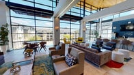 This contemporary single-floor clubhouse is defined by its striking architectural design, most notably the expansive floor-to-ceiling curtain wall windows that flood the interior with natural light and provide panoramic views of the surrounding community and outdoor pool area. The soaring double-height ceilings amplify the sense of openness, with structural beams and integrated linear lighting adding a refined industrial touch. A warm wood-paneled ceiling accent in one wing of the space introduces natural texture, contrasting beautifully with the clean white walls and light tile flooring throughout.

The main lounge area is thoughtfully arranged with multiple distinct seating groupings, including a large sectional sofa, blue accent chairs, and bold yellow swivel chairs that serve as eye-catching focal points. Vibrant area rugs in deep reds, blues, and golds anchor each seating zone, while a bold teal feature wall adds a lively pop of color near the main entrance. A wall-mounted flat-screen television and cozy sofa create a relaxed media lounge nook. A bar-height counter lined with upholstered stools provides an additional social gathering point. Large-scale abstract artwork punctuates the walls, contributing to the space's modern and energetic ambiance. Overall, the space is designed to encourage community interaction in a stylish, light-filled environment.