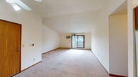 This single-floor residence offers a thoughtfully arranged layout with a variety of functional living spaces. The main living area is notably spacious, featuring vaulted ceilings that add a sense of openness and volume. Neutral-toned carpet flows throughout the living and bedroom areas, complemented by cream-colored walls and warm wood trim. A wall-unit air conditioner and baseboard heating provide year-round climate control, while a sliding glass door floods the living area with natural light and provides outdoor access.

The galley-style kitchen is efficiently designed with warm wood cabinetry extending to the upper walls, a double stainless steel sink, laminate countertops, and white appliances including a refrigerator and electric range. Light tile flooring ties the kitchen together. A ceiling fan is centrally located near the kitchen opening, enhancing airflow throughout the common areas.

The bedrooms are generously sized, each featuring plush carpet, baseboard heating, and windows that frame lush greenery views. Two bathrooms serve the unit—one with a soaking tub and wide vanity, the other with a shower and similarly appointed vanity, both finished with tile flooring and warm wood cabinetry. The overall ambiance is clean, neutral, and move-in ready.