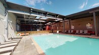 This pool area showcases a striking industrial aesthetic, blending open-air and semi-covered zones into a cohesive amenity space. The centerpiece is a generously sized swimming pool with clear turquoise water, bordered by a wide tiled deck that provides ample room for lounging and circulation. A row of sling-style lounge chairs lines one side of the pool beneath a partially covered overhang supported by steel columns, offering shade while maintaining an open, airy atmosphere under the open sky.

The surrounding structure features exposed steel beams, visible ductwork, and brick walls, lending the space an industrial character. String lights are suspended overhead, suggesting the area is well-suited for evening gatherings. Wooden privacy panels with steel framing delineate sections of the deck, creating defined zones within the larger space.

Adjacent to the pool deck is an indoor area visible through large openings, equipped with a projection screen, pendant lighting, a ventilated cooking station, and colorful accent walls — indicating a flexible event or gathering space. Outdoor seating with round tables and chairs is distributed throughout the deck, providing additional areas for socializing. Overall, the space combines functional amenities with a vibrant, loft-inspired design.