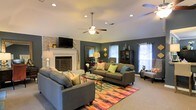 This single-floor home offers a thoughtfully designed living space that balances comfort and style. The centerpiece of the living room is a classic brick fireplace painted in white, paired with a wall-mounted television, giving the space both charm and modern functionality. Deep slate-gray walls create a bold yet cozy backdrop, complemented by abundant natural light streaming through multiple windows dressed with white blinds and sheer curtains. A ceiling fan with integrated lighting anchors the room overhead, enhancing airflow and comfort. The living area is generously sized, comfortably accommodating two large gray sofas accented with colorful throw pillows in shades of teal, green, and coral. A vibrant, multicolored area rug grounds the seating arrangement and adds a lively focal point to the room. A glass-top coffee table sits at the center, while a decorative mirror, cabinet, and accent lamps contribute to the room's layered, curated aesthetic. Recessed lighting throughout the ceiling provides additional illumination. The carpeted flooring adds warmth underfoot, and the overall ambiance of the space is welcoming, relaxed, and full of personality.