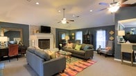 This single-floor home offers a thoughtfully designed living space that balances comfort and style. The centerpiece of the living room is a classic brick fireplace painted in white, paired with a wall-mounted television, giving the space both charm and modern functionality. Deep slate-gray walls create a bold yet cozy backdrop, complemented by abundant natural light streaming through multiple windows dressed with white blinds and sheer curtains. A ceiling fan with integrated lighting anchors the room overhead, enhancing airflow and comfort. The living area is generously sized, comfortably accommodating two large gray sofas accented with colorful throw pillows in shades of teal, green, and coral. A vibrant, multicolored area rug grounds the seating arrangement and adds a lively focal point to the room. A glass-top coffee table sits at the center, while a decorative mirror, cabinet, and accent lamps contribute to the room's layered, curated aesthetic. Recessed lighting throughout the ceiling provides additional illumination. The carpeted flooring adds warmth underfoot, and the overall ambiance of the space is welcoming, relaxed, and full of personality.