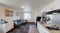 This single-floor studio unit offers a thoughtfully designed open-plan layout that combines the living, dining, and kitchen areas into one cohesive space. The living area features a large L-shaped sectional sofa set against a painted concrete block wall adorned with nautical-themed artwork, complemented by a wall-mounted flat-screen television. A window with sheer white curtains allows natural light to filter into the room, enhancing its bright and airy feel. Wood-look flooring runs throughout the main area, accented by patterned area rugs that add visual warmth and texture.

The kitchenette is efficiently organized along one wall, featuring white cabinetry with granite-look countertops, a stainless steel sink, a full-size refrigerator, a microwave, and open shelving for storage. A small dining table with chairs is tucked nearby, providing a dedicated space for meals. A white dresser offers additional storage within the living area.

The private bathroom is clean and modern in design, with a two-tone wall treatment in white and taupe, wood-look tile flooring, a white vanity, a toilet, and a glass-enclosed walk-in shower. An illuminated LED mirror serves as both a functional and stylish focal point. The overall ambiance of the unit is casual, coastal-inspired, and practical.