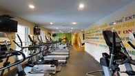 This single-floor fitness center is designed to inspire and support an active lifestyle. The space is generously sized, with rows of modern cardio equipment — including treadmills, ellipticals, and stationary bikes — arranged for ease of movement and accessibility. Recessed lighting illuminates the room evenly, creating a bright and welcoming workout environment. The flooring features a durable dark rubberized surface, ideal for high-traffic fitness use. One of the most striking design elements is the bold motivational mural along the main wall, featuring colorful typography with uplifting fitness phrases in vibrant yellow, green, and blue tones, adding energy and personality to the space. Accent walls in yellow and green further enhance the lively atmosphere. Additional strength and functional training equipment, including medicine balls and benches, are visible toward the rear of the room, providing a well-rounded workout area. Wall-mounted televisions are positioned for viewing during cardio sessions. A window near the entrance brings in a touch of natural light, complementing the recessed overhead lighting throughout. The overall ambiance is motivating, modern, and purposefully designed to encourage fitness and movement.