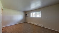 This single-floor unit offers a straightforward, functional layout across several well-defined rooms. The living area features warm, neutral-toned carpet and a wall-mounted air conditioning unit, complemented by a wide window fitted with blinds that allows natural light to fill the space. A baseboard heating system runs along the walls, providing consistent warmth throughout.

The kitchen is a standout area, showcasing dark espresso-toned cabinetry with sleek bar-pull hardware, granite-style countertops with a stainless steel sink, and light wood-look flooring. A ceiling fan with a light fixture overhead adds both comfort and practicality to this space.

The bedrooms are generously sized, each finished with plush carpet and neutral wall tones. Multiple windows provide natural light and outdoor views. One area includes two doors — one leading to a closet and one to a utility space housing a water heater, reflecting a thoughtful use of available space.

The bathroom features a clean, all-white tub and shower enclosure with chrome fixtures, maintaining a bright and tidy aesthetic. Throughout the unit, clean white trim, textured ceilings, and a consistent neutral color palette create a cohesive and livable atmosphere well-suited for everyday comfort.