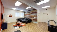 This single-floor space combines contemporary design elements with rustic touches to create a welcoming and visually dynamic environment. The centerpiece of the main lounge area is a striking floor-to-ceiling accent wall composed of reclaimed wood planks in an eclectic mix of natural, painted, and weathered finishes — including tones of red, teal, brown, and grey — providing a bold focal point that adds warmth and character to the space. The lounge is furnished with a large black leather sectional sofa paired with a matching ottoman, arranged around a simple square coffee table. A patterned area rug in red, white, and black grounds the seating area and adds a pop of color to the light hardwood flooring that runs throughout. The drop ceiling features recessed panel lighting that evenly illuminates the room, complementing the natural light admitted through the windows along one wall. The clean white walls and open layout lend an airy, uncluttered feel to the space. Additional doors visible from the lounge suggest access to adjoining rooms or private areas, making this a versatile space well-suited for use as a common area, collaborative lounge, or casual meeting environment.