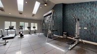 This fitness center is a comprehensively outfitted workout facility designed to support a wide range of exercise disciplines. The space features soaring vaulted ceilings with skylights that flood the interior with natural light, complemented by recessed lighting and a ceiling fan to maintain a comfortable environment. The flooring throughout consists of durable rubber sport tiles, providing cushioning and traction ideal for high-intensity activity.

A standout feature is the TreadWall — a motorized vertical climbing wall — set against a boldly textured teal accent wall that adds a striking visual element to the space. Adjacent to it is a cable machine and weight bench, rounding out the strength-training zone. A full-length mirror with Peloton branding reflects the room and visually expands the space, while dual wall-mounted flat-screen televisions offer entertainment throughout workouts.

The cardio section is lined with large windows that provide views of the outdoor pool area, creating a bright and motivating atmosphere. Multiple treadmills and stationary spin bikes are arranged along this windowed wall. An open stretching and functional training area features parallel bars and wall-mounted televisions, along with a water fountain station for convenience. The overall ambiance is modern, energetic, and purposefully designed to offer a full-spectrum fitness experience.