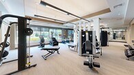 Located at 80 Mill St in Hanover, Massachusetts, this professionally designed fitness center offers a generous, open single-floor layout dedicated entirely to exercise and wellness. The space is thoughtfully organized into distinct zones, providing a seamless workout experience for users of all fitness levels.

The cardio section lines the perimeter near large windows, allowing natural light to flood in while offering pleasant outdoor views of the surrounding greenery. This area features a row of modern treadmills, stair climbers, ellipticals, and stationary bikes, all equipped with digital displays.

Toward the interior, a well-appointed strength training zone includes a multi-functional cable machine, a power rack with a flat bench, medicine ball storage, and a variety of free weights. A decorative column with circular mirror accents serves as an architectural focal point, dividing the space with visual interest while maintaining an open feel.

The design aesthetic is contemporary and clean, with neutral-toned carpet flooring, a coffered ceiling with integrated linear lighting, and warm gold accent panels that add a refined touch. Full-length mirrors along key walls enhance the sense of depth and openness throughout. The overall ambiance is bright, modern, and energizing, creating an inviting environment well-suited for a full range of fitness activities.