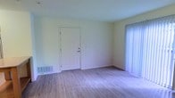 This single-floor space offers a clean and functional layout designed for comfortable living. The main living area is immediately welcoming, featuring light-toned hardwood-style flooring that runs throughout and lends a warm, cohesive feel to the interior. Neutral, pale green-white walls create a bright and airy atmosphere, maximizing the effect of natural light that filters in through the large sliding glass door fitted with vertical blinds, offering easy access to an outdoor area. The front entry door is a classic six-panel white door, adding a traditional touch to the space. A floor-level HVAC vent is positioned along the wall, ensuring efficient climate control throughout. The room benefits from an open, uncluttered layout that provides flexibility for a variety of furniture arrangements. The overall ambiance is simple and practical, making it well-suited for those seeking a straightforward, move-in ready space. The light flooring, pale walls, and generous natural light work together to create an environment that feels open and easy to personalize.