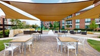 Located at 1502 Partnership Way in Houston, Texas, this thoughtfully designed outdoor communal space serves as a central gathering hub within a multi-building residential community. The courtyard is anchored by a striking pavilion structure featuring natural stone columns and a peaked canopy roof, equipped with ceiling fans and built-in grilling stations — ideal for outdoor entertaining and casual dining.

Extending from the pavilion, large tan shade sails stretch across a broad paver patio, providing ample sun protection over multiple dining tables and white accent chairs arranged throughout. The patterned brick and concrete paver ground surface adds visual texture and defines the various activity zones across the courtyard.

A gently winding concrete walkway connects the different areas of the space, guiding visitors through manicured green lawns and thoughtfully arranged landscaping featuring trees, shrubs, and natural stone accent walls. Rattan lounge seating clusters offer relaxed gathering spots throughout the grounds.

At the far end of the courtyard, a colorful children's playground provides a dedicated recreational area for younger visitors. The surrounding multi-story brick buildings with large windows and private balconies frame the courtyard, creating an enclosed yet open-air atmosphere. The overall design balances functionality and aesthetics, fostering a welcoming outdoor environment for the community.