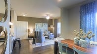 This single-floor residence features an open-concept layout that seamlessly connects the dining and living areas, creating a cohesive and functional living space. The flooring throughout is a warm-toned hardwood, adding a sense of continuity and elegance across the space.

The dining area is positioned toward the front and accommodates a solid wood table paired with contemporary seating, providing a practical yet stylish setting for meals. The living area beyond is anchored by a large abstract artwork in muted blues and greens, serving as a focal point against the taupe-toned walls. Comfortable upholstered seating is arranged around a central area rug, fostering an inviting atmosphere.

A ceiling fan with a light fixture provides overhead illumination in the living space, complemented by floor and table lamps that add warm, layered lighting throughout. A window dressed with patterned drapes allows natural light to filter into the room. Crown molding adds a refined architectural detail, while a solid entry door with a modern finish contributes to the overall polished aesthetic of the space.