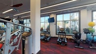 This modern fitness center is designed to support a full spectrum of workout routines in a bright, energizing environment. The main workout floor is expansive, featuring bold red patterned carpeting that adds a dynamic visual energy to the space. Large floor-to-ceiling windows line the perimeter, flooding the interior with natural light and offering views of palm trees and the surrounding outdoor landscape.

The cardio zone is well-appointed with a row of treadmills, ellipticals, and stationary bikes, each positioned to take advantage of the outdoor views. Multiple wall-mounted flat-screen televisions are strategically placed throughout for entertainment during workouts. The strength training area includes a comprehensive selection of Hoist selectorized machines, adjustable benches, a cable system, and a full dumbbell rack ranging across a wide variety of weights.

A dedicated free weight and functional training corner is stocked with medicine balls, kettlebells, resistance tools, and a punching bag, catering to diverse training styles. Ceiling fans complement the linear LED lighting overhead, contributing to a comfortable workout atmosphere.

Adjacent to the main floor is a separate studio room with warm hardwood flooring, mirrored walls, and a large mounted display screen, ideal for group fitness classes or personal training sessions. Full-length mirrors throughout the facility enhance the sense of space and support proper form during exercise.