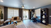 This thoughtfully designed single-floor residence blends contemporary style with functional living across a well-organized layout. The main living area is bright and welcoming, featuring wood-look flooring, a ceiling fan, recessed lighting, and large windows with plantation shutters and floor-length draping curtains that invite abundant natural light. A sliding glass door opens directly to a private outdoor balcony, extending the living space outdoors. A striking accent wall in deep charcoal anchors the living room, which accommodates dual seating areas and a wall-mounted television. Adjacent to the living space is an intimate dining nook framed by dark cabinetry display cases, a marble-topped round table, and a bold framed art piece — a standout design moment. The kitchen is well-appointed with grey wood-tone cabinetry, white quartz countertops, a tile backsplash, black appliances, and a peninsula with bar seating beneath pendant lighting. The primary bedroom offers plush carpet, a ceiling fan, natural light through shuttered windows, and a serene, restful ambiance. A second bedroom features a dramatic upholstered headboard wall and a ceiling fan. The bathroom includes a walk-in shower with glass doors, wood-look tile flooring, and a dark vanity with white countertop.