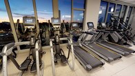 This well-appointed fitness center offers a premium workout experience, combining high-end equipment with an inspiring urban backdrop. The space is lined with expansive floor-to-ceiling windows that frame breathtaking panoramic views of the city skyline, filling the room with natural light during the day and offering a dazzling cityscape at dusk and beyond. The facility is outfitted with a comprehensive selection of professional-grade Matrix cardio equipment, including multiple treadmills and elliptical cross-trainers, each unit featuring integrated digital display screens for tracking performance metrics. The light-toned flooring contributes to a clean, spacious atmosphere that feels both functional and inviting. The layout is thoughtfully arranged to maximize usable floor space while ensuring each piece of equipment has ample room for safe and comfortable use. The combination of modern machine design, abundant glass, and the ever-present city views creates an energizing ambiance that motivates users throughout their workout. This fitness center is well-suited for those seeking a high-quality exercise environment with an elevated urban aesthetic.