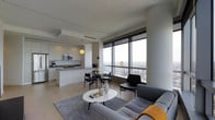This sophisticated single-floor residence exemplifies contemporary urban living, characterized by a clean, minimalist aesthetic and an open-concept design that seamlessly integrates the living, dining, and kitchen areas. Floor-to-ceiling windows dominate the living space, flooding the interior with natural light and framing breathtaking panoramic views of the city skyline stretching far into the distance — a standout feature that immediately commands attention upon entry.

The living area is anchored by a plush gray sectional sofa, a round white coffee table, and a soft area rug, creating a comfortable and stylish lounge space. Warm pendant lighting accents complement the neutral white and gray color palette found throughout. The dining area, positioned adjacent to the windows, features a compact glass-top table with sleek black chairs, making the most of the spectacular views.

The kitchen is thoughtfully designed with flat-panel white cabinetry, a gray stone countertop, an undermount sink, and stainless steel appliances including a microwave and refrigerator. Light hardwood flooring flows continuously throughout the space, contributing to the cohesive, airy feel. The overall ambiance is polished and modern, ideal for those who appreciate refined, urban design with a strong connection to the city beyond.