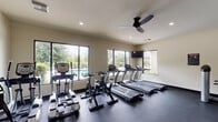 This fitness center offers a comprehensive workout environment designed to support a wide range of exercise routines. The space is organized into distinct functional zones, with cardio equipment positioned along the windowed perimeter and a dedicated strength training area occupying the interior. Large windows line the walls, flooding the room with natural light and providing views of an outdoor pool and surrounding landscaping, creating an energizing atmosphere during workouts.

The cardio section includes multiple treadmills, stationary bikes, and an elliptical trainer, all positioned to take advantage of the outdoor views. A wall-mounted flat-screen television is installed above the treadmill row, offering entertainment during cardio sessions. Ceiling fans are mounted throughout, ensuring proper air circulation across the space.

The strength training area features a Smith machine, a cable cross machine, a weight bench, and a full rack of dumbbells in graduated sizes. A large floor-to-ceiling mirror spans one wall of the strength zone, visually expanding the space and allowing users to monitor their form during exercises.

The flooring throughout is dark rubber gym matting, providing durability and traction. Recessed lighting supplements the natural light, keeping the space well-lit at all times. The neutral-toned walls and clean layout contribute to a functional yet welcoming fitness environment.