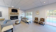 This single-floor clubhouse offers a welcoming and functional space designed for community use and social gatherings. The layout flows naturally between two distinct areas, each serving a unique purpose.

The lounge area is anchored by a striking floor-to-ceiling stacked stone fireplace fitted with a gas insert and topped with a rustic wood mantel, above which a wall-mounted flat-screen television is displayed. Comfortable seating arrangements, including leather sofas, accent chairs, and ottomans, create an inviting atmosphere. A sliding glass door opens to views of an outdoor sports court, filling the room with natural light.

Adjacent to the lounge, a spacious dining area accommodates multiple high-top tables with leather upholstered chairs, all oriented toward large windows that frame the outdoor courts. This area transitions seamlessly into a well-equipped kitchenette featuring warm maple cabinetry, dark granite countertops, a built-in microwave, a full-size refrigerator, and a stainless steel sink. A breakfast bar with bar stools adds additional seating options.

Throughout the space, wood-look flooring in the kitchen and entry areas contrasts with neutral carpeting in the lounge and dining zones. The drop ceiling with recessed and panel lighting ensures consistent, functional illumination throughout the entire clubhouse.