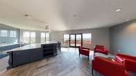 This single-floor communal space combines a well-appointed kitchen and a comfortable lounge area into one cohesive, open layout. The kitchen is outfitted with navy blue cabinetry, a large central island with quartz countertops and built-in open shelving, pendant lighting, and a full suite of stainless steel appliances including a refrigerator, built-in oven, and beverage cooler. A full-height glossy subway tile backsplash in a cool slate-blue tone adds a polished, contemporary touch, while multiple windows flood the area with natural light and frame outdoor views.

The lounge area flows seamlessly from the kitchen, anchored by bold red leather armchairs arranged around a low center table. A wall-mounted flat-screen television sits atop a built-in navy blue credenza, creating a functional media nook. Recessed lighting and smooth gray walls contribute to the clean, modern atmosphere throughout.

Underfoot, a striking herringbone-patterned tile floor in a warm wood-look finish runs throughout the entire space, adding visual depth and warmth. French doors along one wall open directly to an outdoor terrace, where red seating and sweeping elevated views extend the usable space beyond the interior. An additional private room, accessible via a glass-paneled door, rounds out the layout.