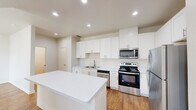 Located along N IH-35 in Austin, TX, this two-story residence offers 2 bedrooms, 3 bathrooms, and a well-organized layout spread across two floors. The main level features an open-plan kitchen and living area finished with warm hardwood-style flooring and recessed lighting throughout. The kitchen is outfitted with white shaker cabinetry, white quartz countertops, a central island, stainless steel appliances including a refrigerator, range, microwave, and dishwasher, and a double-basin sink — combining a clean, contemporary aesthetic with practical functionality. The living area flows naturally from the kitchen, creating a cohesive and bright common space. Upstairs, the spacious primary bedroom offers generous square footage with plush carpeting, a ceiling fan, and a dedicated closet. The second bedroom is also carpeted and features a window allowing natural light. Both floors are served by staircases, and the residence includes three full bathrooms — one featuring a tiled soaking tub with brushed gold fixtures, and another with a glass-enclosed walk-in shower, wood-style flooring, and a white vanity. The overall palette of whites and neutrals creates a clean, modern ambiance throughout.